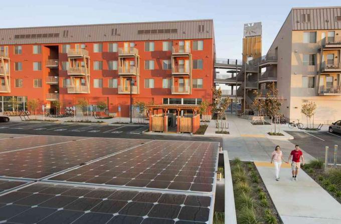 Dua orang berjalan di dekat gedung hijau dan panel surya, dalam komunitas Zero Net Energy