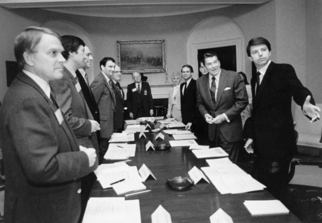 Gambar hitam putih Ronald Reagan dan beberapa pria berjas di sekitar meja konferensi yang panjang